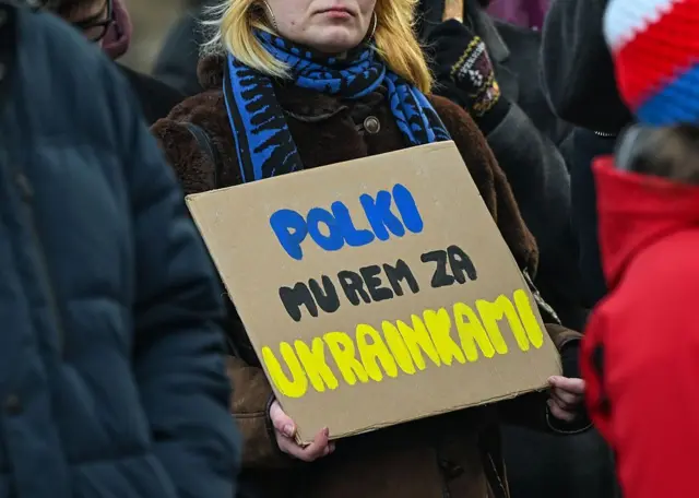 Жінка тримає картонний плакат із написом польською синьо‑жовтими літерами: "Польки стоять горою за українок". Навколо – інші учасники заходу, що стоять у натовпі.