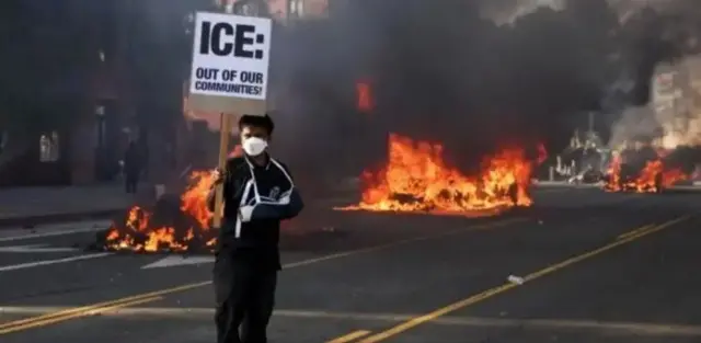 Людина стоїть на фоні пожежі з табличкою "ІСЕ геть"