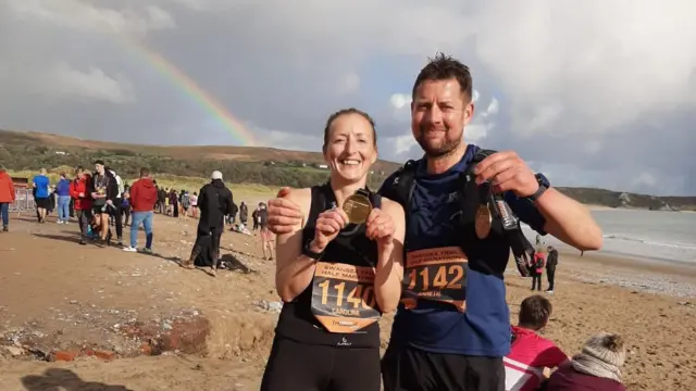 Gareth e Caroline, com roupas de corrida, na praia de Swansea (País de Gales). Eles estão sorrindo e segurando medalhas de corrida