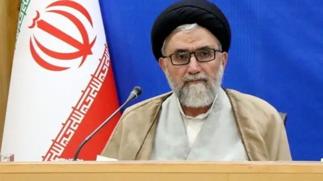 اسماعیل خطیب، در کنار او پرچم جمهوری اسلامی است و پس‌زمینه تصویر رنگ آبی است