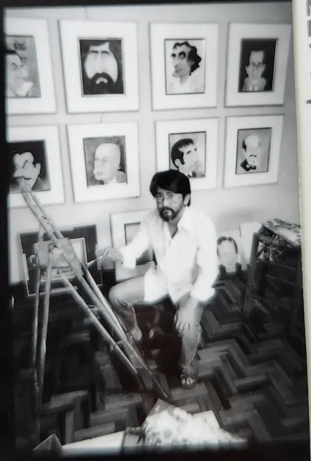 A imagem em preto e branco mostra Cláudio Seto, um homem barbudo e de óculos, agachado em um ateliê ou sala de trabalho, cercado por quadros com caricaturas pendurados na parede. À frente dele há um cavalete de madeira, enquanto outros retratos apoiados no chão e móveis aparecem ao redor. O ambiente tem piso de madeira em padrão geométrico e aspecto de arquivo fotográfico antigo.