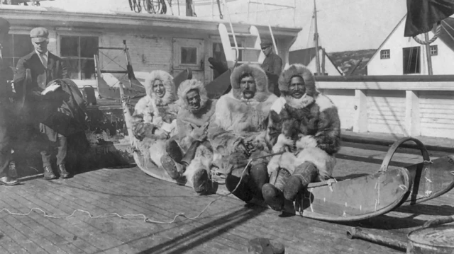 Photo en noir et blanc. Matthew Henson en compagnie de trois autres explorateur. Ils sont tous les quatre assis sur une luge disposée sur la plage avant du bateau. Le groupe porte une tenue d'hiver. 