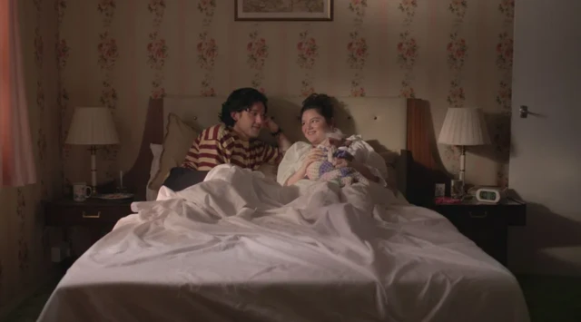 Un hombre (i) y una mujer conversan sonrientes en una cama. 