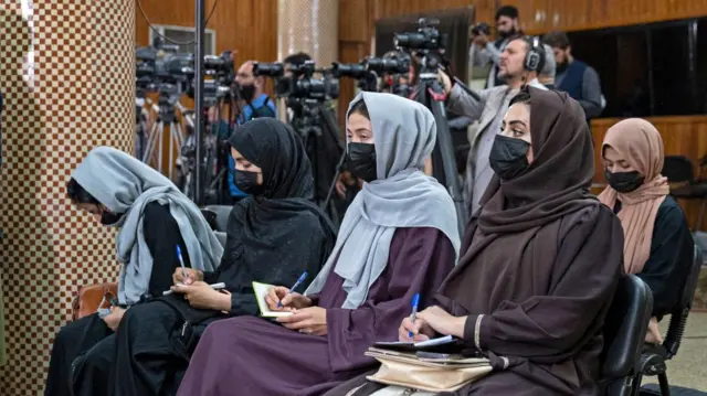 A line of seated women wearing burkas and face masks hold pens and notebooks surrounded by cameras