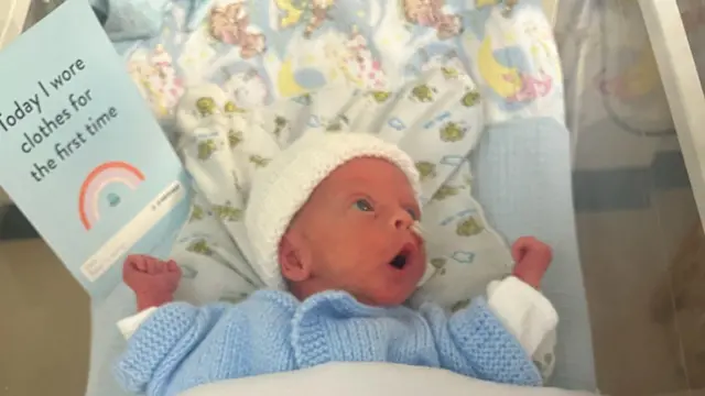Jude en la cuna de la unidad neonatal. Sostiene una tarjeta que dice "Hoy estrené ropa". Lleva un cárdigan de punto azul y un gorro de punto blanco. Mira hacia un lado de la cuna, boquiabierto, con expresión de sorpresa o emoción.