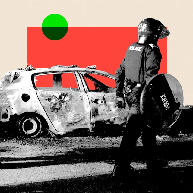 Black and white image of a police officer in Hartlepool wearing riot gear walking past a burnt out car