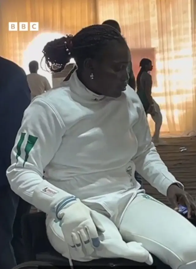 Aanu Kumuyi wọ asọ oke ati sokoto alawọ funfun, o wọ ibọwọ si ọwọ kan, o joko lori aga irinsẹ onirin, to si han pe wọn ti ge ẹsẹ rẹ kan
