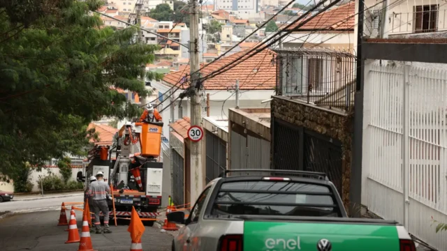 Carro da Enel, agência de energia, e funcionários da empresa, em frente a um poste de energia