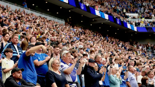 Setki kibiców Evertonu, ubranych w niebiesko‑białe barwy, stoją na stadionie Hill Dickinson, unosząc telefony podczas meczu towarzyskiego Evertonu z AS Romą w 2025 r.