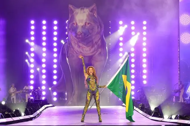 Shakira no palco em Copacabana, segurando uma bandeira do Brasil com uma estátua de um lobo gigante ao fundo