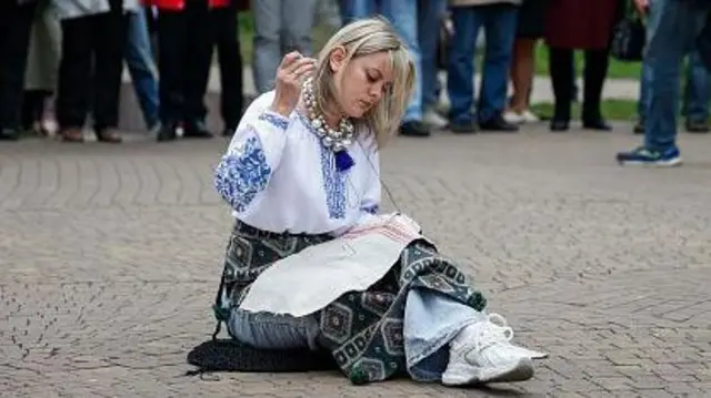 Дівчина у вишиванці сидить на бруківці і вишиває , позаду неї стоять люди.