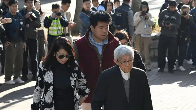 サングラスをかけ、黒と白のスーツを着た黒髪の女性と、えんじ色のジャケットを着た背の高い黒髪の青年と、白髪でめがねをかけスーツ姿の男性が歩いている。3人のすぐ背後にめがねをかけた女性がいるが、顔などは見えない。さらにその後ろには、報道陣がカメラを構えて3人を撮影している