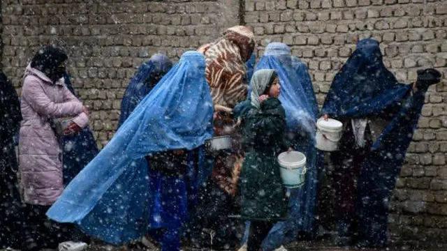 تصویر چند زن و یک دختر در صف توزیع کمک. این زنان ظرف‌های پلاستکی در پست دارند و در زیر بارش برف و هوای سرد منتظر توزیع کمک هستند. 