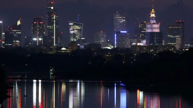 Nocny widok budynków Warszawy, na pierwszym planie światła miasta odbijające się w Wiśle.