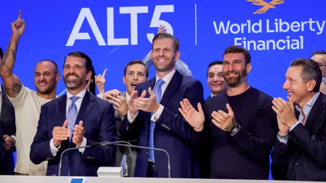 Eric and Donald Trump in dark blue suits and light blue ties clap hands at the Nasdaq stock exchange surrounded by other executives from World Liberty Financial in August 2025