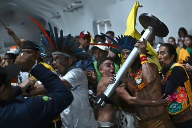 мужчина с обнаженным торсом, с металлической стойкой ограждения в руках и в головном уборе из синих перьев пытается силой пробиться сквозь толпу, его удерживают другие мужчины