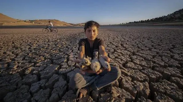 پسربچه‌ای در کف یک روخانه کاملا خشک نشسته و ماهی‌های خشک شده را در دست گرفته به سمت دوربین عکاس نشان می‌دهد