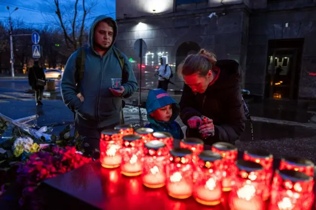 Bir çocuk, bir kadın ve erkek kırmızı cam kaselerde mum yakıyor.