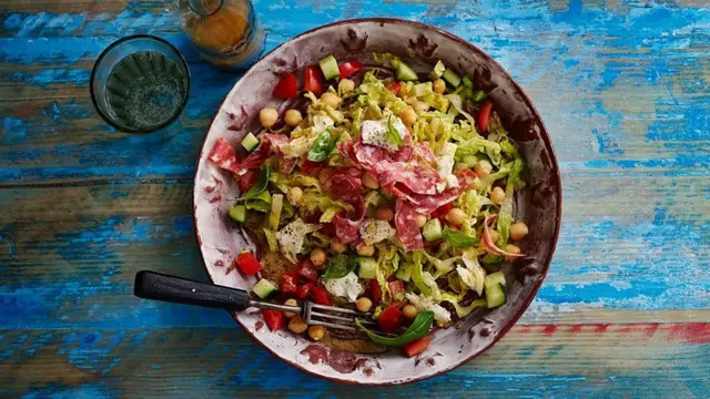 Miska z kolorową sałatką zawierającą ciecierzycę, ogórek, pomidory, sałatę i plastry mięsa, ustawiona na matowym niebieskim stole.