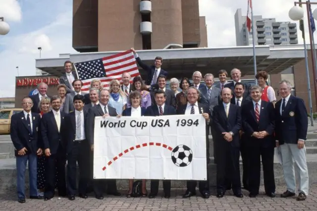 Grupo de pessoas perfiladas exibe a bandeira da Copa do Mundo de 1994