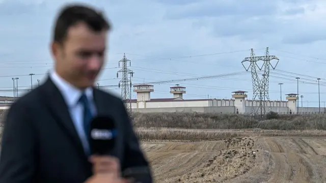 Bir gazeteci elinde mikrofonla ayakta duruyor. Arkada ise Silivri'deki Marmara Cezaevi görülüyor. 