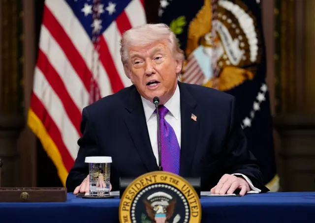 Трамп сидить за столом під час офіційного виступу або пресконференції. На столі розміщено табличку з печаткою "Президент Сполучених Штатів", а позаду – прапори США та елементи офіційної державної символіки.