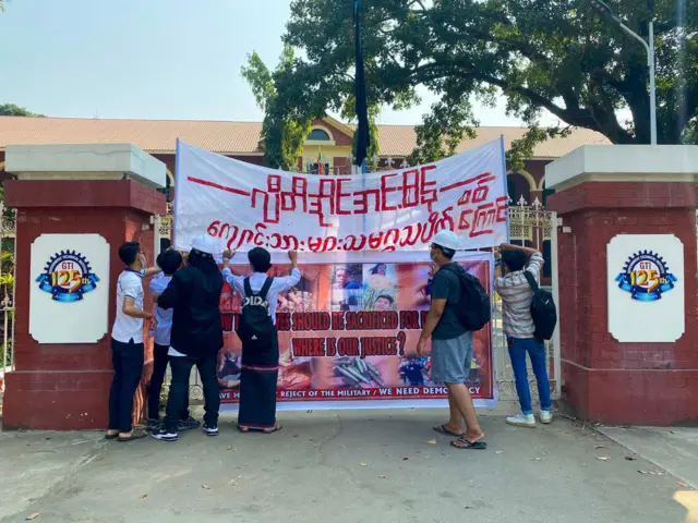 ဂျီတီအိုင်အင်းစိန် ကျောင်းသားများသမဂ္ဂသပိတ်စစ်ကြောင်း အနီရောင်စာလုံးပါ အဝတ်စနဲ့ လုပ်ထားတဲ့ ဆိုင်းဘုတ်ကို ကျောင်းဂိတ်ဝမှာ တပ်ဆင်နေတဲ့ လူငယ် ၆ ဦး 