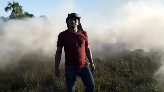 Homem com chapéu e camisa vermelha posa com fumaça e vegetação de cerrado ao fungo.