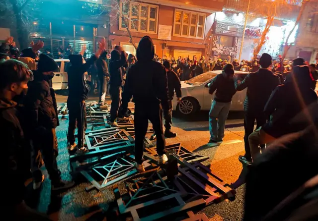 در تصویر، جمعیتی از مردم در خیابان‌های تهران برای اعتراض تجمع کرده‌اند. افراد نزدیک به هم ایستاده‌اند و برخی پلاکارد یا دست‌هایشان را بالا گرفته‌اند. فضا شهری است و ساختمان‌ها در پس‌زمینه دیده می‌شود.  تهران، ایران – ۸ ژانویه ۲۰۲۶