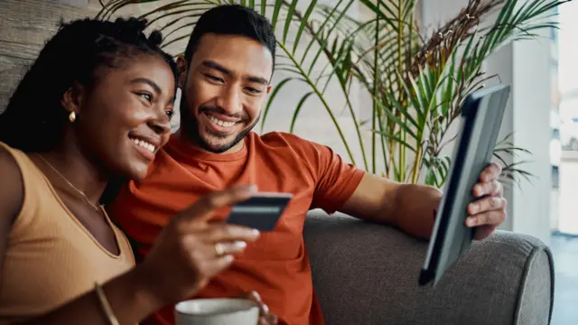 Couple sitting on a sofa with a woman holding a bank card and a man holding a tablet computer. Both are smiling.