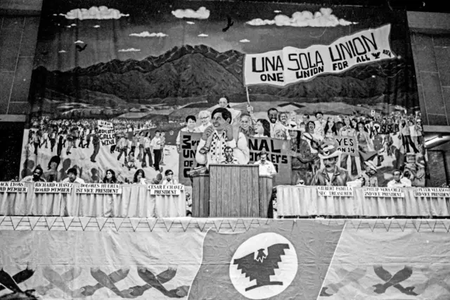 Bajo una enorme pancarta, el representante de los trabajadores agrícolas de InterHarvest, Armando Ruiz, habla durante la convención de United Farm Workers (UFW), Fresno, California, 1975. En el escenario detrás de él están, de izquierda a derecha, los miembros de la junta de UFW Mack Lyons y Richard Chavez, la 1.ª Vicepresidenta Dolores Huerta, el Presidente Cesar Chavez (1927 - 1993), el Secretario Tesorero Gilbert Padilla, el 2.º Vicepresidente Philip Vera Cruz y el 3.º Vicepresidente Peter Velasco. (Foto de Cathy Murphy/Getty Images)