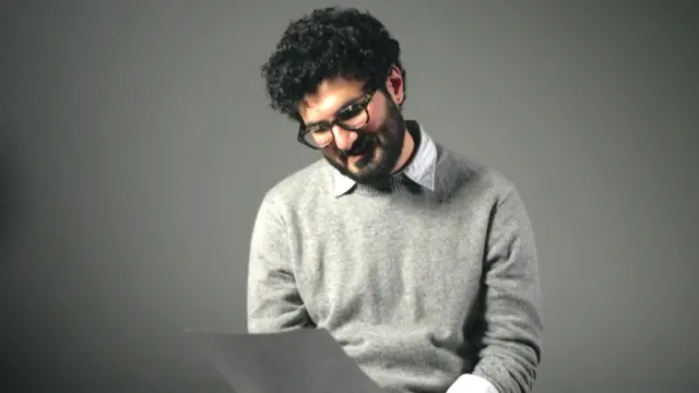 Homem com cabelo ondulado escuro, barba e bigode, usando óculos e uma blusa cinza, olha para uma folha de papel