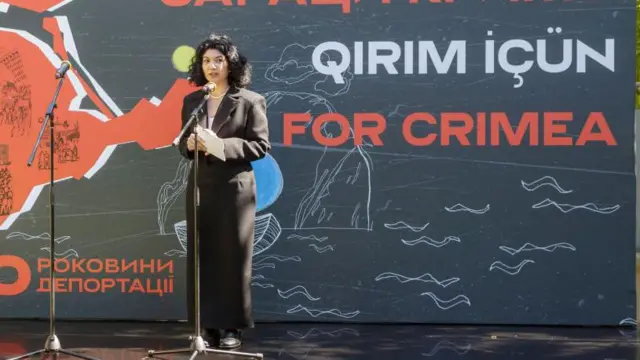 Tamila Taşeva bir Kırım etkinliğinde konuşurken, arkasında Qırım İçün yazıyor.
