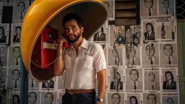 Wagner Moura aparece no papel de um professor em O Agente Secreto. O ator fala em um telefone público vermelho, sob uma cabine amarela, com expressão séria e concentrada. Ao fundo, cartazes políticos antigos cobrem a parede, sugerindo um contexto de tensão política e ambientação histórica.