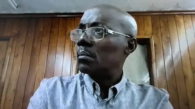 Gérard Ntakirutimana, asa nk'uraramye, yambaye indorerwamo, ishati igaragara nk'ubururu bwerurutse, inyuma ye haboneka urukuta rw'imbaho. Ifoto yakuwe muri videwo y'urukiko iburanisha ririmo kuba, ku wa kabiri, ku itariki ya 19 Ugushyingo (11) mu mwaka wa 2024.