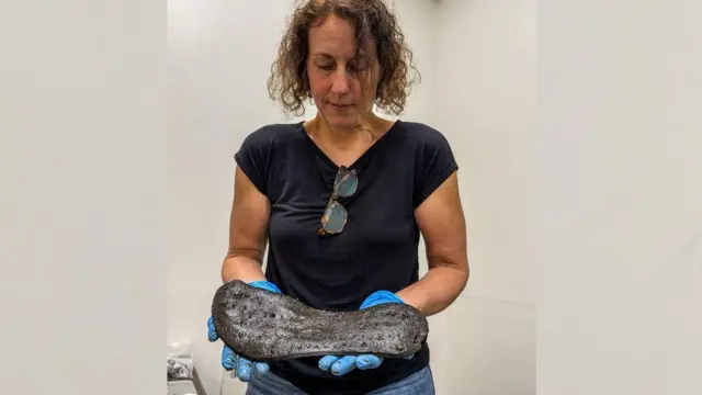 La Dra. Elizabeth Greene lleva una camiseta negra, guantes azules y gafas de sol colgando de la parte superior. En sus manos lleva la suela de un zapato negro grande.
