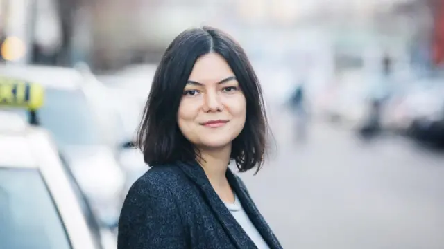 Yaka siyah ceket beyaz tşört ile yol kenarında durmuş kameraya bakıyor, onun dıiında her şey flu. 