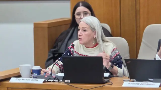 Kay Mason-Billig está sentada tras una mesa en una reunión del consejo. Tiene una computadora portátil y un micrófono frente a ella. Lleva un jersey navideño color crema arriba y estampados rojos, azules y grises abajo.
