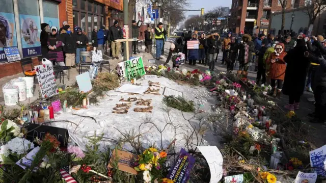 道路の一角で雪の周りを花束やカードが囲み、十字架が立てられ、大勢がそれを囲んでいる