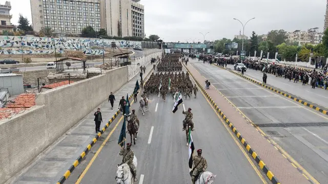 Askeri geçit töreninde en önde atlı askerler var. Arkada yaya şekilde askerler geçit töreni yapıyor. Fotoğrafı önden ve havadan çekilmiş şekilde görüyoruz