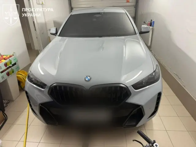 Сірий автомобіль BMW стоїть у невеликому гаражі або підсобному приміщенні. Навколо — побутові речі та обладнання, а машина виглядає чистою й неушкодженою.