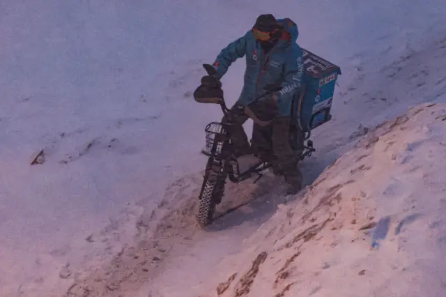 Доставщик еды пробирается на велосипеде по узкой полоске между сугробами