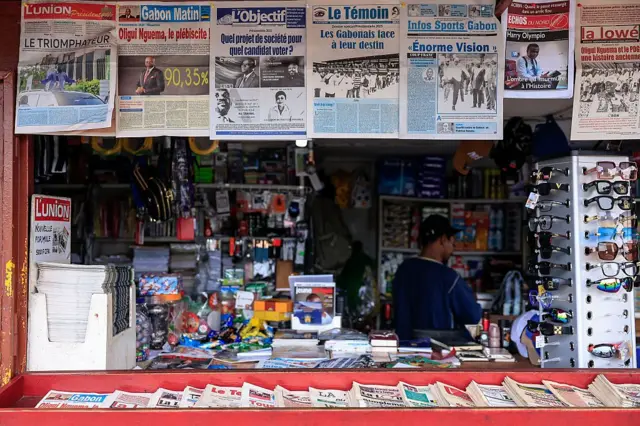 Une kiosque avec différents titres de journaux devant, des lunettes exposées à côté et d'autres accessoirs à l'intérieur. Une femme portant un haut bleu est également visible à l'intérieur de la kiosque.