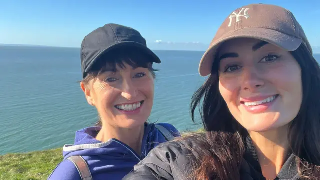 Lauren Macpherson aparece ao lado da mãe em uma costa ensolarada. Lauren (à direita) usa um boné marrom e sorri para a câmera. A mãe também usa um boné, tem franja castanha, olhos castanhos e sorri para a câmera. Ela veste um casaco roxo com zíper e está atrás da filha