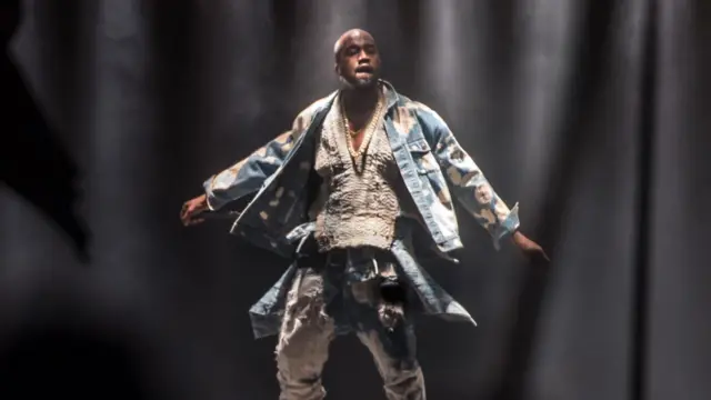 Kanye West stoi na scenie podczas festiwalu Glastonbury w 2015 r., z wyciągniętymi na boki ramionami. 