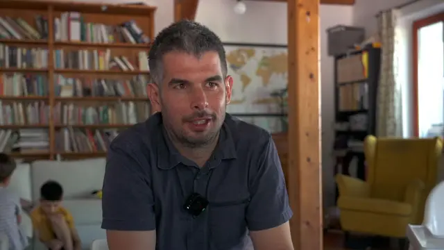 Egy rövid barna hajú férfi látható a kép előterében. Mögötte két kisgyerek játszik a földön ülve egy könyvespolc előtt.
