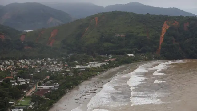 Foto aérea mostra praia e encostas com deslizamentos