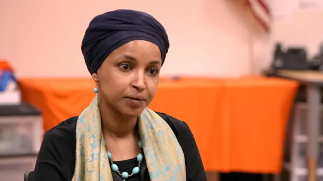 Ilhan Omar apfunze igitambara cirabura mu mutwe, igishora kw'izosi n'impuzu yirabura, yambaye n'ishapure ry'ibara rya turquoise