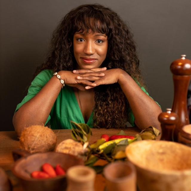 Mulher negra posa em uma mesa cheia de elementos culinários, como moedor de pimenta, coco seco e pimentas 