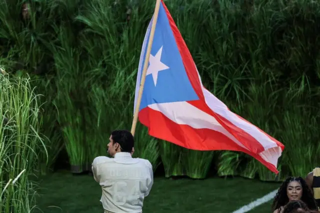 サトウキビ畑を模した舞台の中で、「オカシオ　64」と背中に描かれたクリーム色のアメフトジャージを着たバッド・バニーさんが、紅白の横しまと水色の三角、三角の中に白い星がひとつのプエルトリコの旗を掲げている